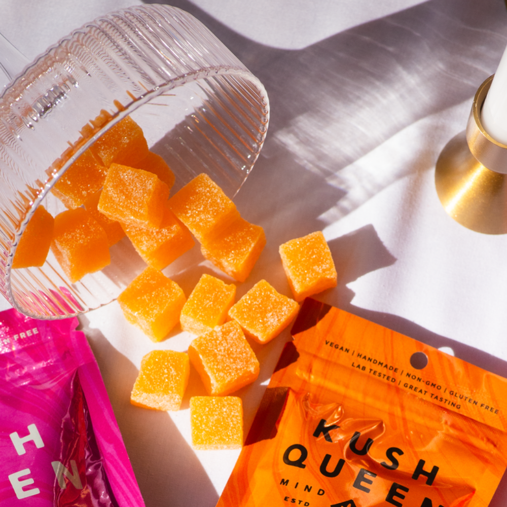 Kush Queen Tangerine THC and CBD gummies spilling out of a glass dish with Kush Queen Tangerine THC and CBD packaging in the background next to it.